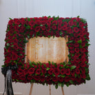 Red rose memorial standing frame — dense red roses and Italian ruscus around a parchment prayer scroll with cross, mounted on a wooden easel | Ecoroses Los Angeles