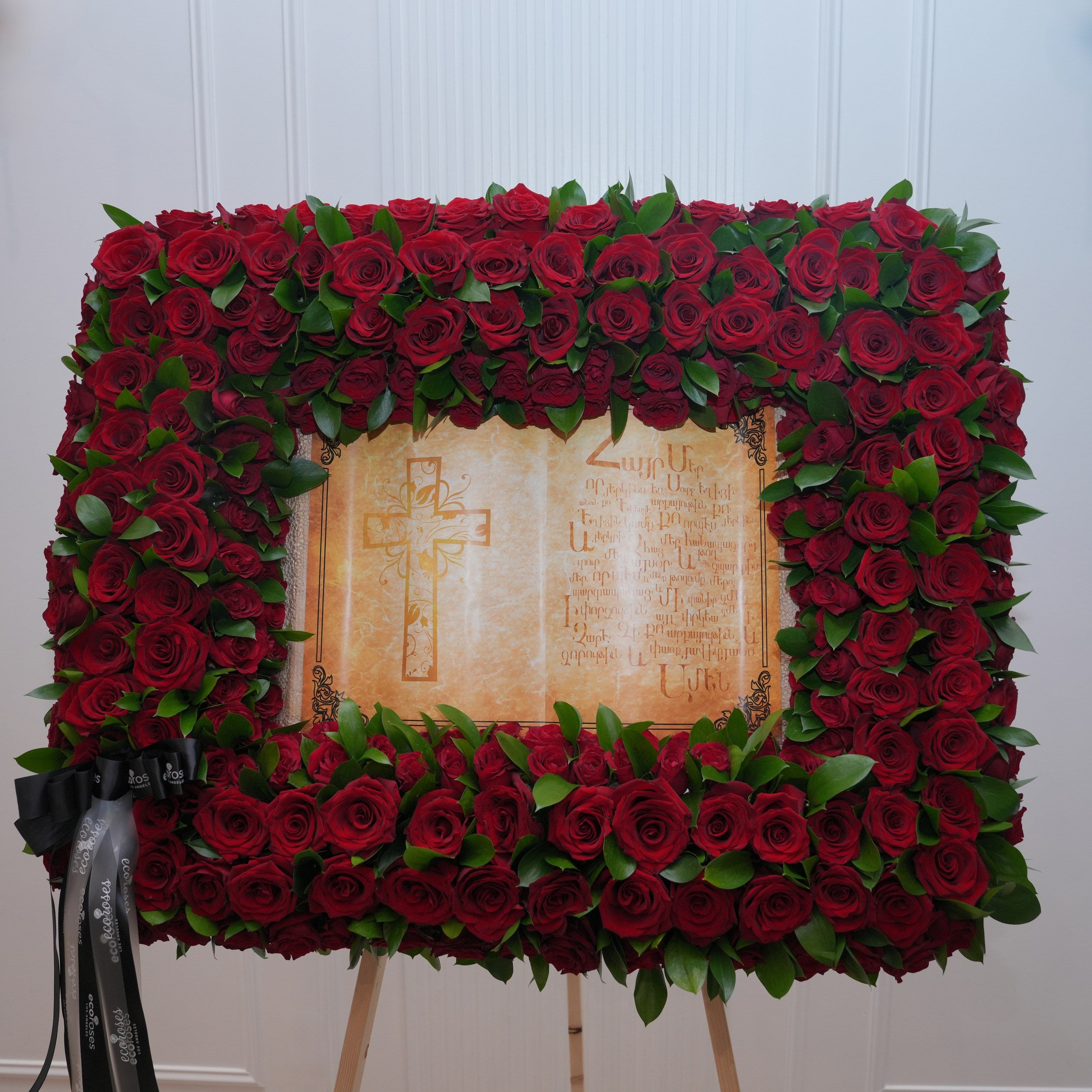 Red rose memorial standing frame — dense red roses and Italian ruscus around a parchment prayer scroll with cross, mounted on a wooden easel | Ecoroses Los Angeles