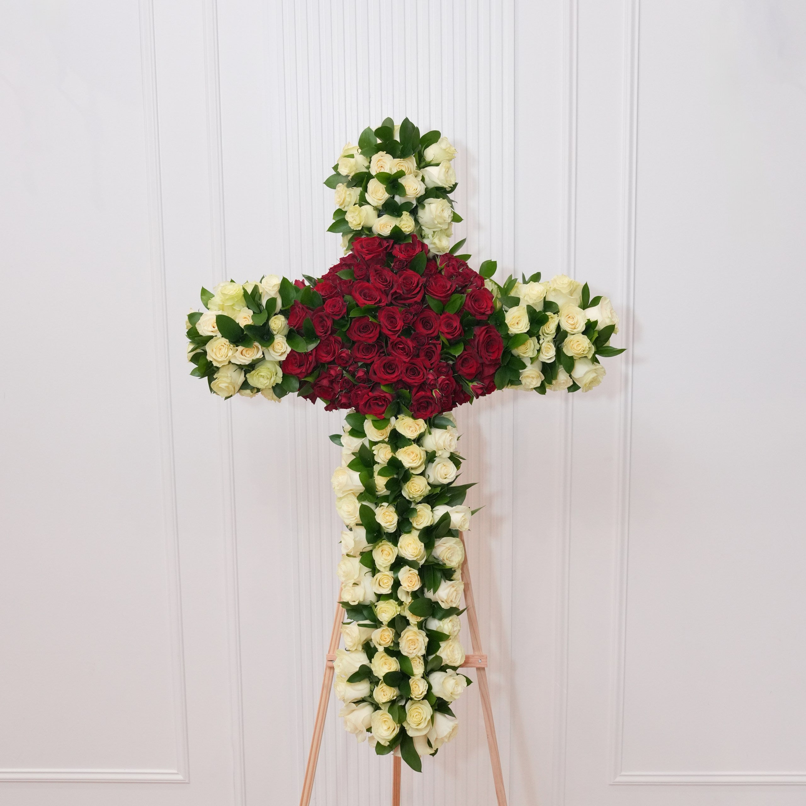 White rose standing cross — white roses along the arms and base with a red rose cluster at the center crossing, backed by Italian ruscus foliage on a wooden easel | Ecoroses Los Angeles